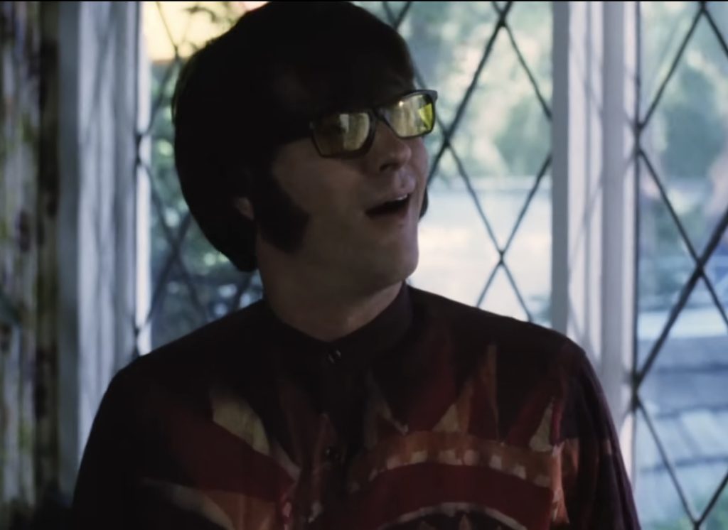 Greg Kean as a 1966 Brian Wilson in Summer Dreams: The Story of The Beach Boys. His character is wearing glasses with yellow tinted lenses and a brown shirt with a tie dye style sun.